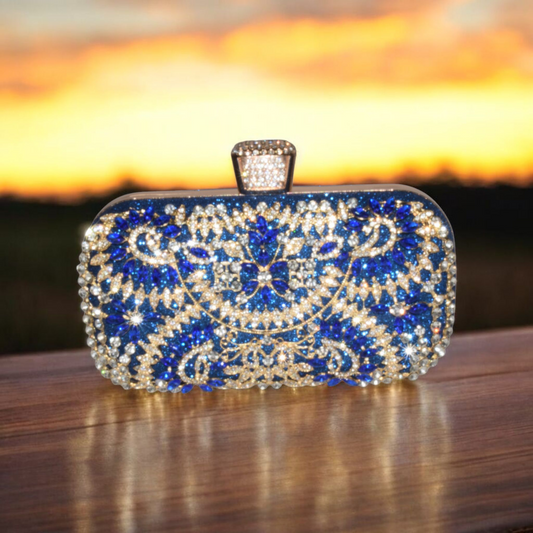 A rhinestone embellished blue and gold rectangle clutch with a chain strap, displayed against a sunset background.