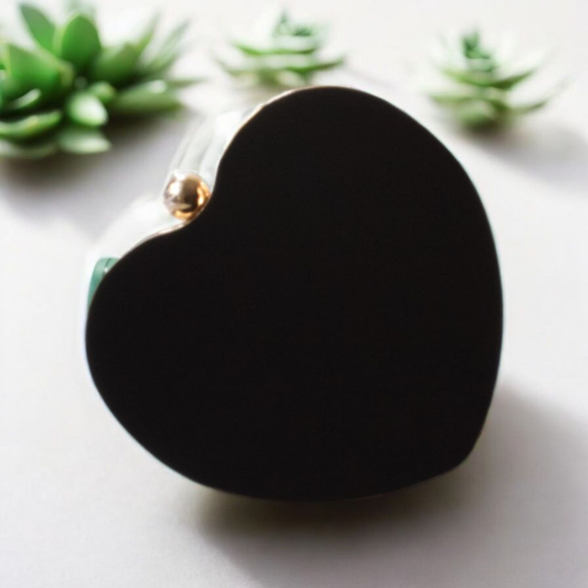Black heart-shaped object with a gold accent on a light surface with green plants in the background